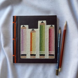 May include: A brown notebook with a colorful pencil design on the cover. The pencils are purple, orange, green, and pink. The notebook has orange stitching on the spine.
