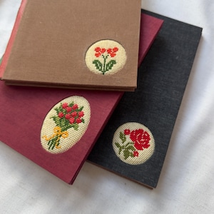 May include: A stack of three small notebooks with embroidered floral designs. The top notebook is brown, the middle is burgundy, and the bottom is dark gray. Each notebook features a different cross-stitched flower design in a circular or oval patch.