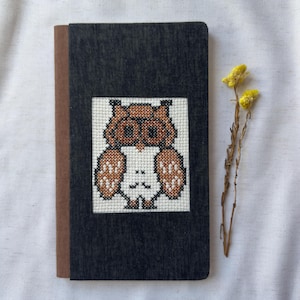May include: A black notebook with a brown border and a cross-stitch design of a brown owl on the front.