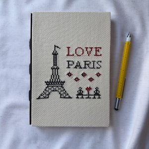May include: A white canvas notebook with black stitching and a black and white embroidered image of the Eiffel Tower. The text "LOVE PARIS" is embroidered in red on the front of the notebook.