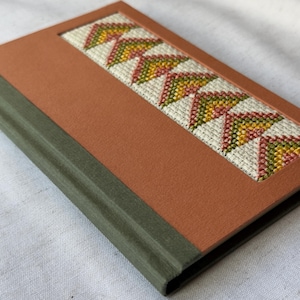 May include: A small, orange-brown journal with a green spine. The front cover features a decorative panel with a stitched design of green, yellow, and pink triangles.