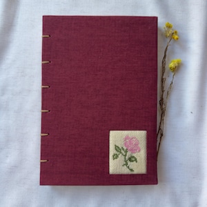 Op de afbeelding: Een bordeauxrood notitieboek met handgestikte binding. Een kleine, vierkante kruissteekpatch met een roze roos en groene bladeren staat op de voorkant. Een takje gele bloemen ligt naast het notitieboek op een wit oppervlak.