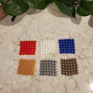 May include: Six small, square, decorative items in various colors: red, white, blue, gold, gray, and brown. Each item has a repeating star-like pattern. The items are arranged on a speckled, light gray surface, with green leaves in the background.