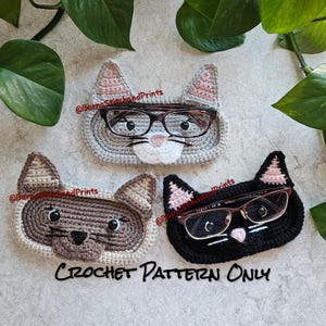 May include: Three crocheted cat-shaped eyeglass holders. The holders are in gray, brown, and black. Each holder has a pair of glasses resting on top. The text "CROCHET PATTERN ONLY" is printed below the holders.