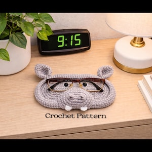 May include: A gray crochet eyeglass holder shaped like a hippo, with brown eyeglasses resting inside. The holder has small ears and white tusks. The text "Crochet Pattern" is visible below the holder.