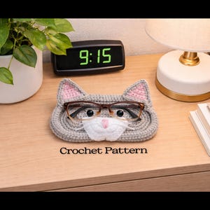 May include: A gray crocheted cat-shaped glasses holder with pink-lined ears and a white nose, holding a pair of eyeglasses. The text "Crochet Pattern" is below the holder. The holder sits on a wooden nightstand next to a lamp and a digital clock.
