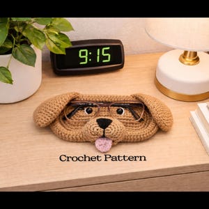 Puede incluir: Un soporte para gafas con forma de perro tejido a ganchillo en tonos marrones, con nariz negra, lengua rosa y gafas en su interior. El texto "Crochet Pattern" es visible. El soporte está sobre una superficie de madera.