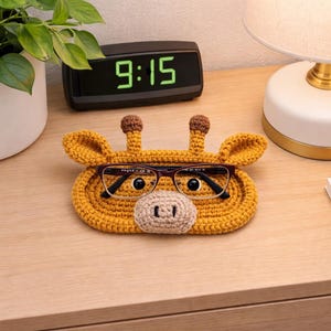 May include: A handcrafted, yellow crochet eyeglass holder shaped like a giraffe, complete with brown ears and horns, and a beige nose. The holder is designed to cradle a pair of eyeglasses. The time on the digital clock is 9:15.