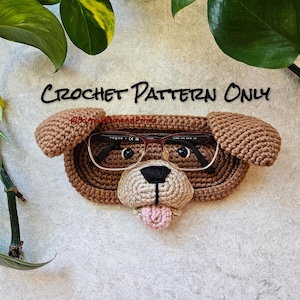 Puede incluir: Un estuche para gafas con forma de perro tejido a ganchillo en tonos marrón, beige y rosa, con un par de gafas dentro. El estuche presenta la cara de un perro con nariz negra, lengua rosa y orejas marrones. El texto "CROCHET PATTERN ONLY" está escrito encima.