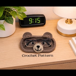 Puede incluir: Un soporte para gafas con forma de oso tejido a ganchillo en tonos marrones, con un hocico color crema. El soporte para gafas está sobre una superficie de madera, con un reloj digital que muestra las 9:15 y el texto "Crochet Pattern".