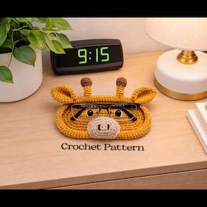 May include: A handmade, yellow crochet giraffe-shaped glasses holder with brown accents. The holder has a beige nose and holds a pair of eyeglasses. The text "Crochet Pattern" is visible below the holder. The holder is on a wooden surface.