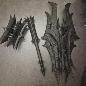 Puede incluir: Un conjunto de armas de fantasía negras, que incluye un hacha de batalla, una lanza y un escudo. El hacha presenta una hoja con púas y un mango detallado. La lanza tiene una punta larga y afilada. El escudo tiene un diseño texturizado complejo con bordes afilados.