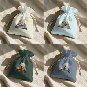 Op de afbeelding: Vier trekkoordzakken in verschillende kleuren: crème, lichtblauw, donkergroen en leisteenblauw. Elke zak heeft een driehoekig ontwerp met een berg en bloemen- of hemelse elementen. De zakken hebben een trekkoordsluiting in natuurlijke kleur.