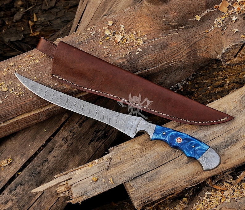 May include: A Damascus steel knife with a long, curved blade and a blue and white handle. The knife rests on a wooden surface next to a brown leather sheath. The handle has a red accent.