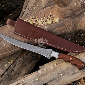 May include: A Damascus steel knife with a curved blade and a wooden handle. The knife has a brown leather sheath. The handle features a red emblem. The knife and sheath are on a wooden surface.
