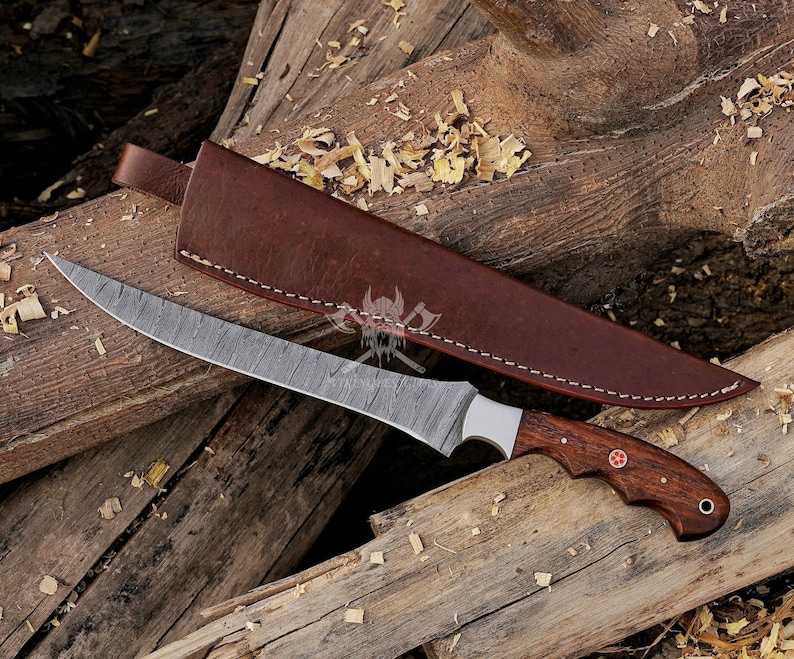 May include: A Damascus steel knife with a curved blade and a wooden handle. The knife has a brown leather sheath. The handle features a red emblem. The knife and sheath are on a wooden surface.