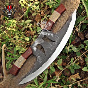 May include: A large, curved blade knife with a dark, textured metal finish. The handle is made of dark wood with a patterned, light-coloured grip. The blade has two decorative cutouts and a sharp edge. The knife is displayed on a tree branch.