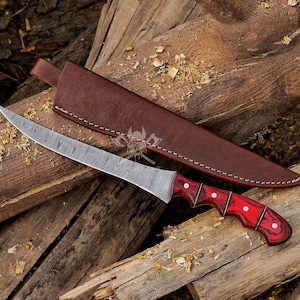 May include: A Damascus steel knife with a curved blade and a red wooden handle with white accents. The knife is paired with a brown leather sheath, resting on a wooden surface. The blade has a unique pattern.