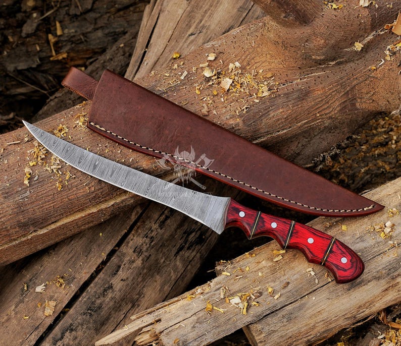 May include: A Damascus steel knife with a curved blade and a red wooden handle with white accents. The knife is paired with a brown leather sheath, resting on a wooden surface. The blade has a unique pattern.