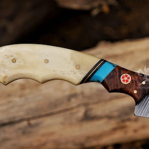 May include: Close-up of a handcrafted knife with a bone handle. The handle features brass rivets, a turquoise inlay, and a dark wood section. The blade has a patterned design. The knife is set against a blurred wooden background.