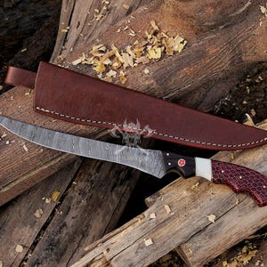 May include: A Damascus steel fillet knife with a brown leather sheath. The knife has a black and red handle with a white accent and a red cross emblem. The blade is curved and has a unique pattern.
