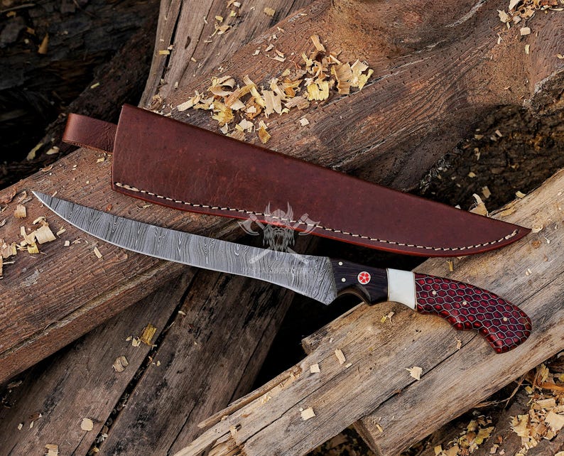 May include: A Damascus steel fillet knife with a brown leather sheath. The knife has a black and red handle with a white accent and a red cross emblem. The blade is curved and has a unique pattern.