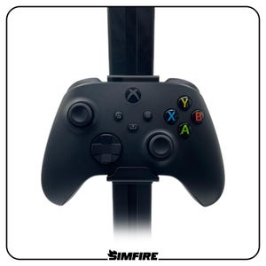 Puede incluir: Un mando de videojuegos inalámbrico negro montado en un soporte vertical. El mando cuenta con un logotipo de Xbox central, dos joysticks analógicos, una cruceta y cuatro botones de acción etiquetados como X, Y, B y A en azul, amarillo, rojo y verde.