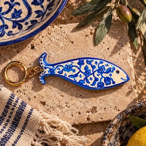 May include: A fish-shaped keychain featuring a cream-colored base with a hand-painted blue floral pattern. The keychain includes a gold-toned metal ring attached to the tail of the fish, which has a smooth, glossy finish and a distinct eye detail.