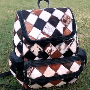 May include: A patchwork backpack with a diamond pattern in shades of brown, black, and white. The backpack has a top handle, two side pockets, and a front pocket, all with black zippers. The bag is made of a textured material.