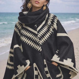 May include: A black and cream-colored poncho with a geometric pattern. The poncho has a high collar and pockets. The model is wearing gold earrings and is standing on a beach.