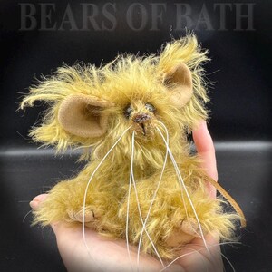 May include: A handmade, plush mouse toy with long, white whiskers and large, tan ears. The mouse has a shaggy, golden-brown fur and is held in a person's hand. The text "BEARS OF BATH" is visible in the background.