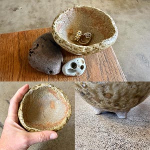 May include: A handmade, textured ceramic bowl with a natural, earthy color palette. The bowl contains gold rings. Two stones, one gray and one white with holes, are next to the bowl. The bowl is on a wooden surface.