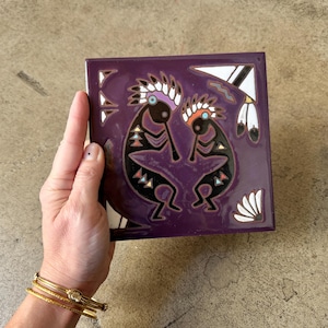 May include: A square, purple tile with a Native American Kokopelli design. The tile features two Kokopelli figures in black, with white, red, and black accents. The tile is held in a hand.