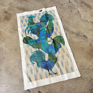May include: A rectangular kitchen towel featuring a repeating pattern of stylized roosters in shades of blue and green. The roosters are set against a beige background with a wheat-like design, framed by a white border.