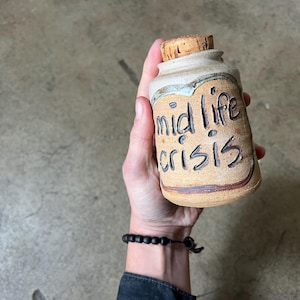 May include: A small, brown ceramic jar with a cork lid. The jar has the words "mid life crisis" written on it in black.
