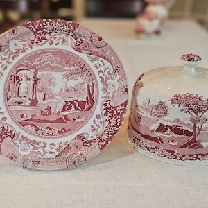 May include: A red and white ceramic cake plate with a dome lid. The plate and lid feature a floral pattern with a central scene of a landscape with trees and buildings.
