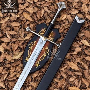 May include: A silver-toned metal sword with a black wrapped handle, a crossguard, and a pommel. The blade features engraved runic text and a star symbol. It is displayed with a black scabbard and a dark brown shield-shaped plaque with gold filigree.