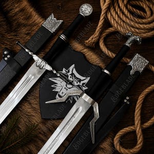 May include: Two steel swords with black grips and silver-toned pommels, one featuring a wolf head design. Both swords include black scabbards with silver metal accents. A black shield-shaped plaque displays a white wolf head emblem in the center.