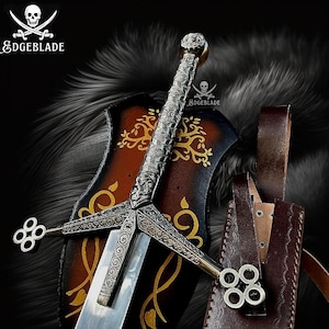 Op de afbeelding: Een zilveren en gouden zwaard met ingewikkelde details, tentoongesteld op een bruin en gouden plaquette met een boomontwerp. Het zwaard heeft een leren schede met een decoratieve riem.