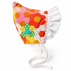 May include: A bonnet featuring a colorful patchwork of hexagonal fabric pieces in shades of red, orange, yellow, green, and pink with various floral and polka dot patterns. The headwear includes a solid white ruffled trim and two white fabric ties.