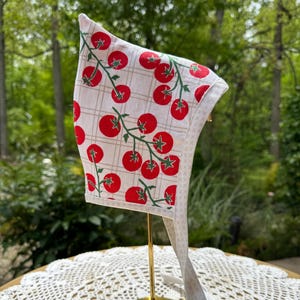 May include: A white bonnet with a red tomato print on a grid pattern. The bonnet has a white trim and ties. The bonnet is displayed on a gold stand against a blurred green background.