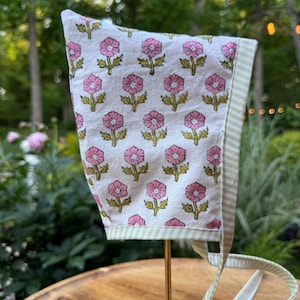 May include: A white fabric bonnet featuring a repeating pattern of pink flowers with green stems and leaves. The bonnet is trimmed with a light green and white striped fabric border and includes matching striped fabric ties for securing the item.