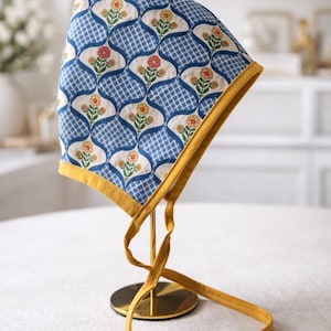 May include: A blue and white patterned bonnet with a floral design, trimmed with yellow fabric. The bonnet is tied with yellow fabric ties and is displayed on a gold stand. The floral pattern includes red and yellow flowers.