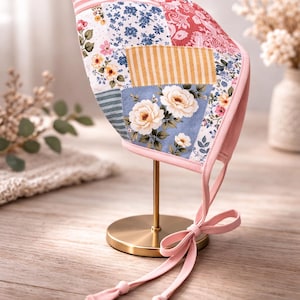 May include: A patchwork bonnet with floral and striped patterns in pink, blue, and yellow. The bonnet is displayed on a gold stand and has pink ties. The bonnet is likely for a baby or child.