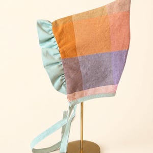 May include: A baby bonnet in a plaid pattern of orange, purple, and pink hues. The bonnet features a light blue ruffled trim and ties. The bonnet is displayed on a gold stand.