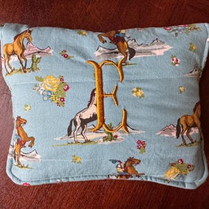May include: A rectangular light blue pillow featuring a repeating pattern of horses, cowboys, mountains, cacti, and floral clusters. The center of the pillow is embroidered with a large, golden-yellow capital letter E in a decorative script font.