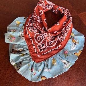 May include: A pet bandana featuring a rust-colored top section with a white paisley pattern and a light blue ruffled bottom section. The light blue fabric is decorated with a repeating print of horses, colorful floral bouquets, and green foliage.