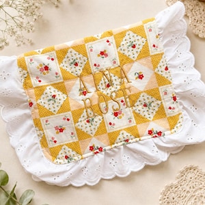 May include: A rectangular quilted fabric pouch featuring a yellow and white diamond pattern with floral motifs and strawberries. The center is embroidered with the text AINA BOST in gold thread, surrounded by a white eyelet lace ruffled border.