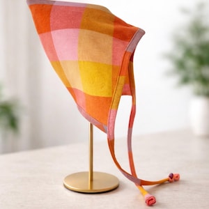 May include: A colorful plaid bonnet in shades of orange, pink, and yellow, displayed on a gold stand. The bonnet has long ties with rolled ends. The fabric pattern is a large check design, creating a cheerful and vibrant look. The bonnet is a baby or toddler accessory.