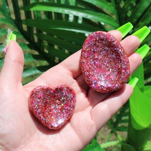 Könnte beinhalten: Zwei kleine, konkave Dekoschalen aus Kunstharz, eine in Herzform und die andere oval. Beide Objekte haben eine dichte, mehrfarbige rosa und rote Glitzeroptik mit reflektierenden metallischen Partikeln, die in die Oberfläche eingearbeitet sind.